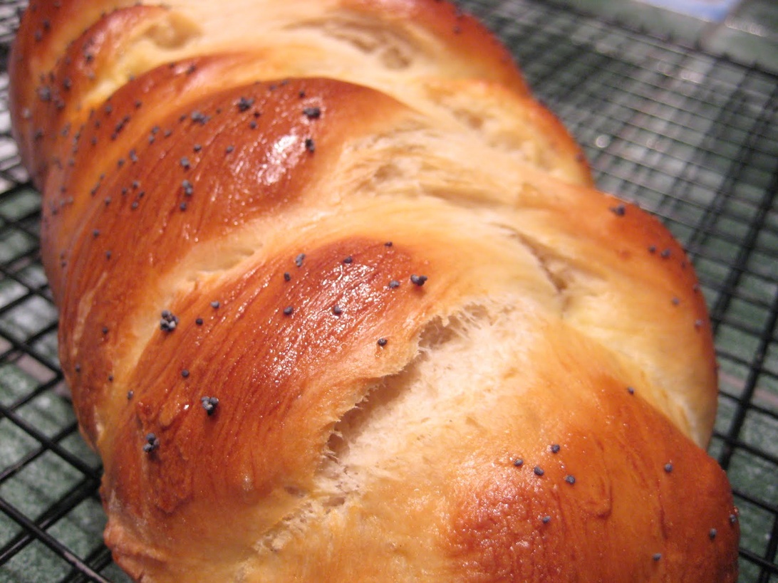 En närbild på ett vackert flätat challahbröd med gyllenbrun yta toppat med vallmofrön, vilande på ett galler. Brödet ser nybakat ut och visar den intrikata flätningstekniken som kräver tålamod och fokus.