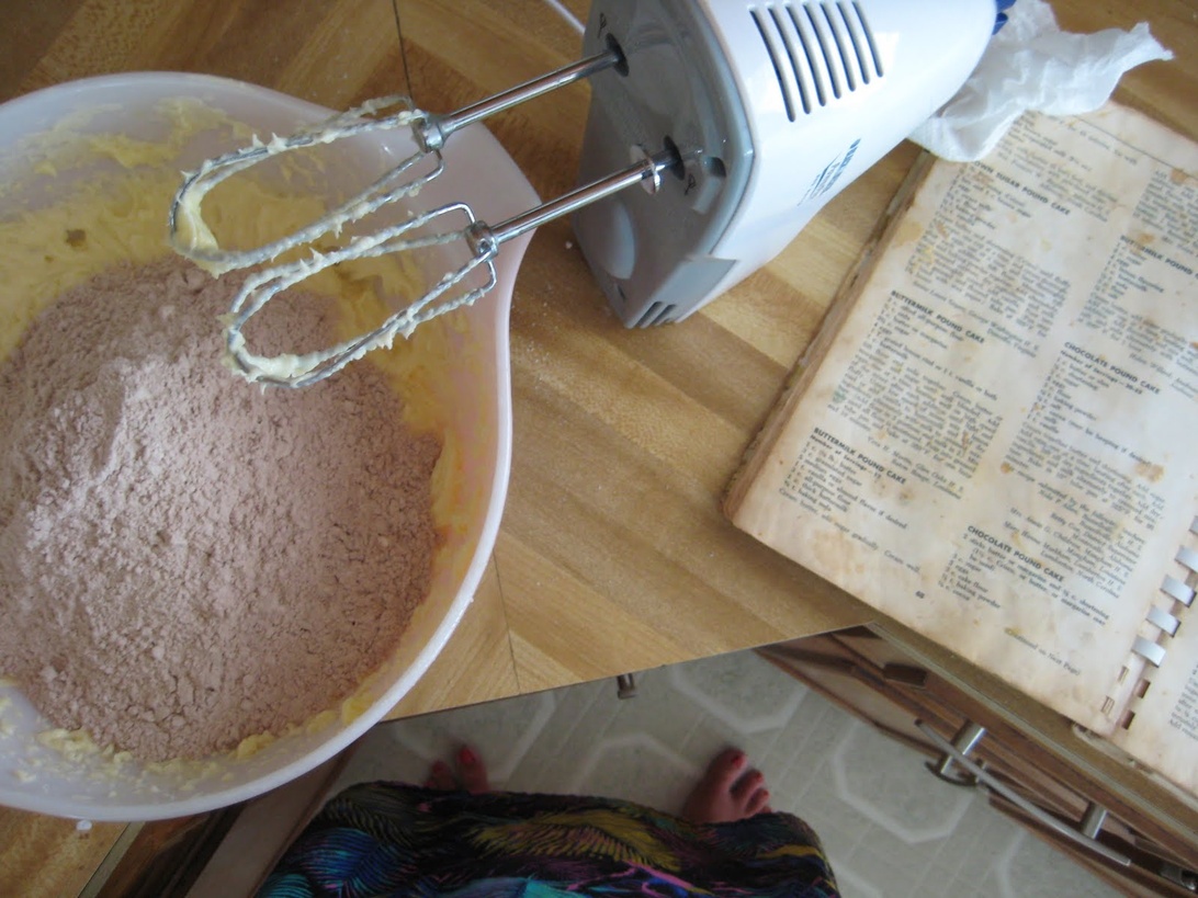 En översiktsbild av en bakscen med en elvisp täckt av smet, en bunke med torra choklad-ingredienser och en öppen, äldre kokbok. Upplägget antyder en hemmabagare som följer ett traditionellt recept, vilket representerar de metodiska, nostalgiska aspekterna av bakning som kan vara terapeutiska.